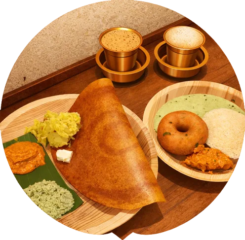 A platter with dosa, chutneys, idli, and a fried vada, accompanied by two cups of filter coffee in stylish brass cups.